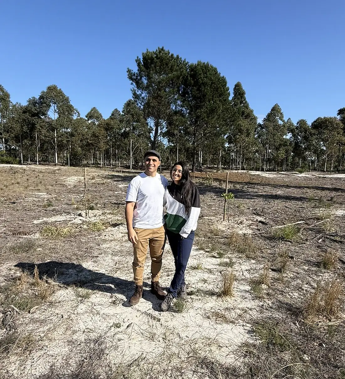 Nacho y Florencia parados en el claro arenoso donde va a ir la casa, con el monte de eucaliptos detrás.